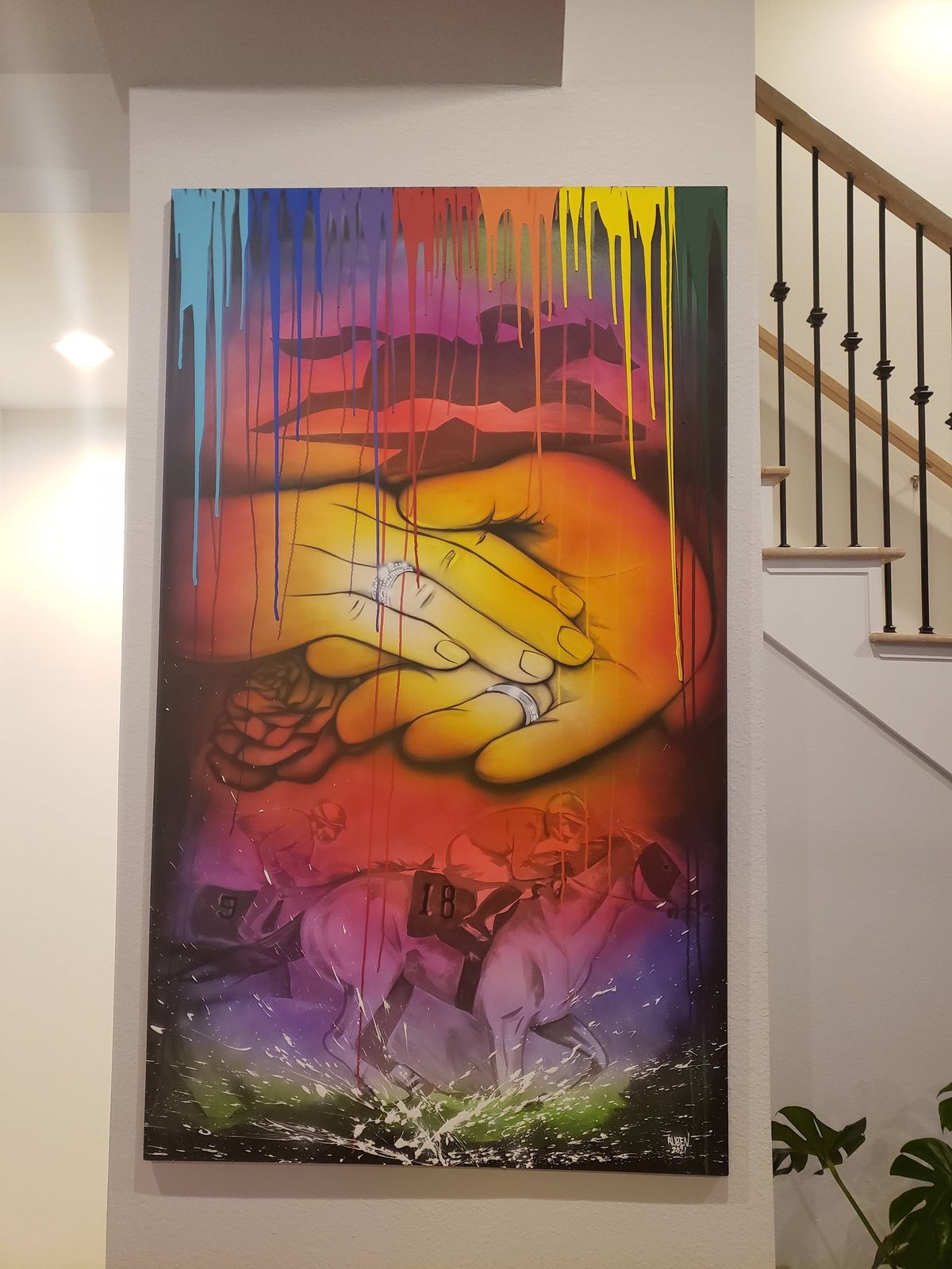 A tall canvas hanging beside a staircase, painted with two joined hands wearing rings, a rose and rainbow paint drips running down from the top.