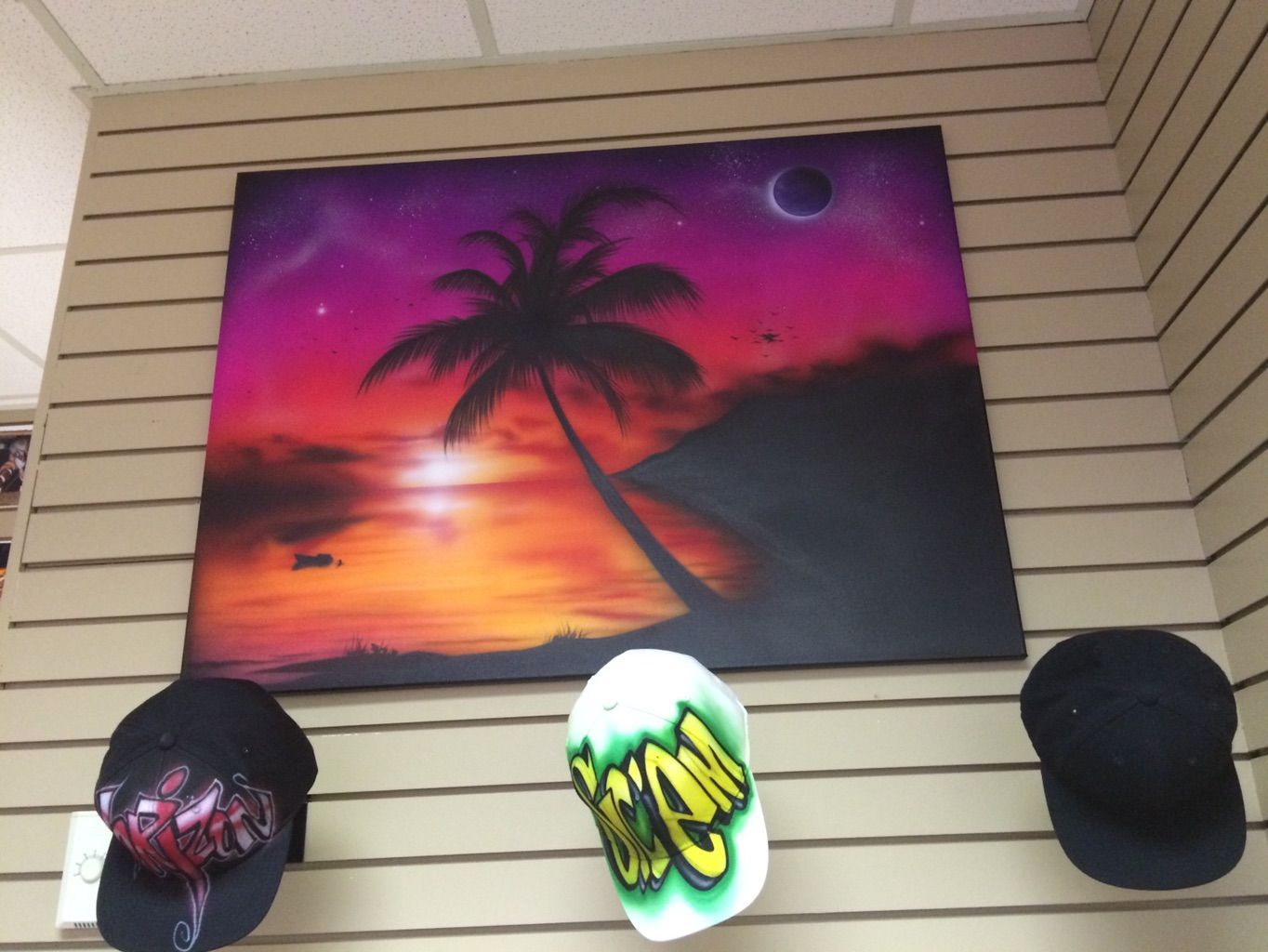 An airbrushed canvas of a palm tree silhouetted against a magenta and orange sunset over water, hung on a slatwall above three caps, two of them airbrushed with graffiti lettering.