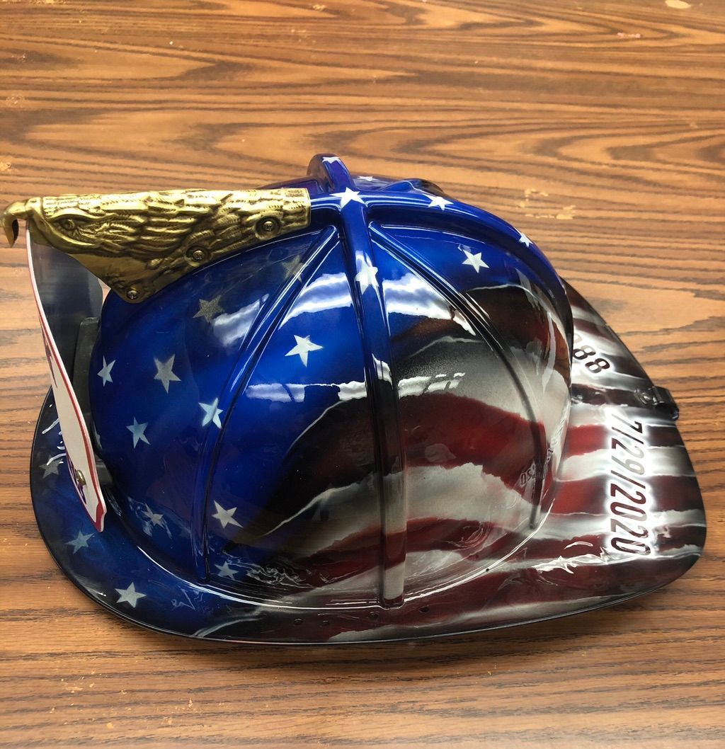 A traditional firefighter helmet with a brass eagle on top, airbrushed with white stars on deep blue and a waving American flag, with service dates painted along the brim.