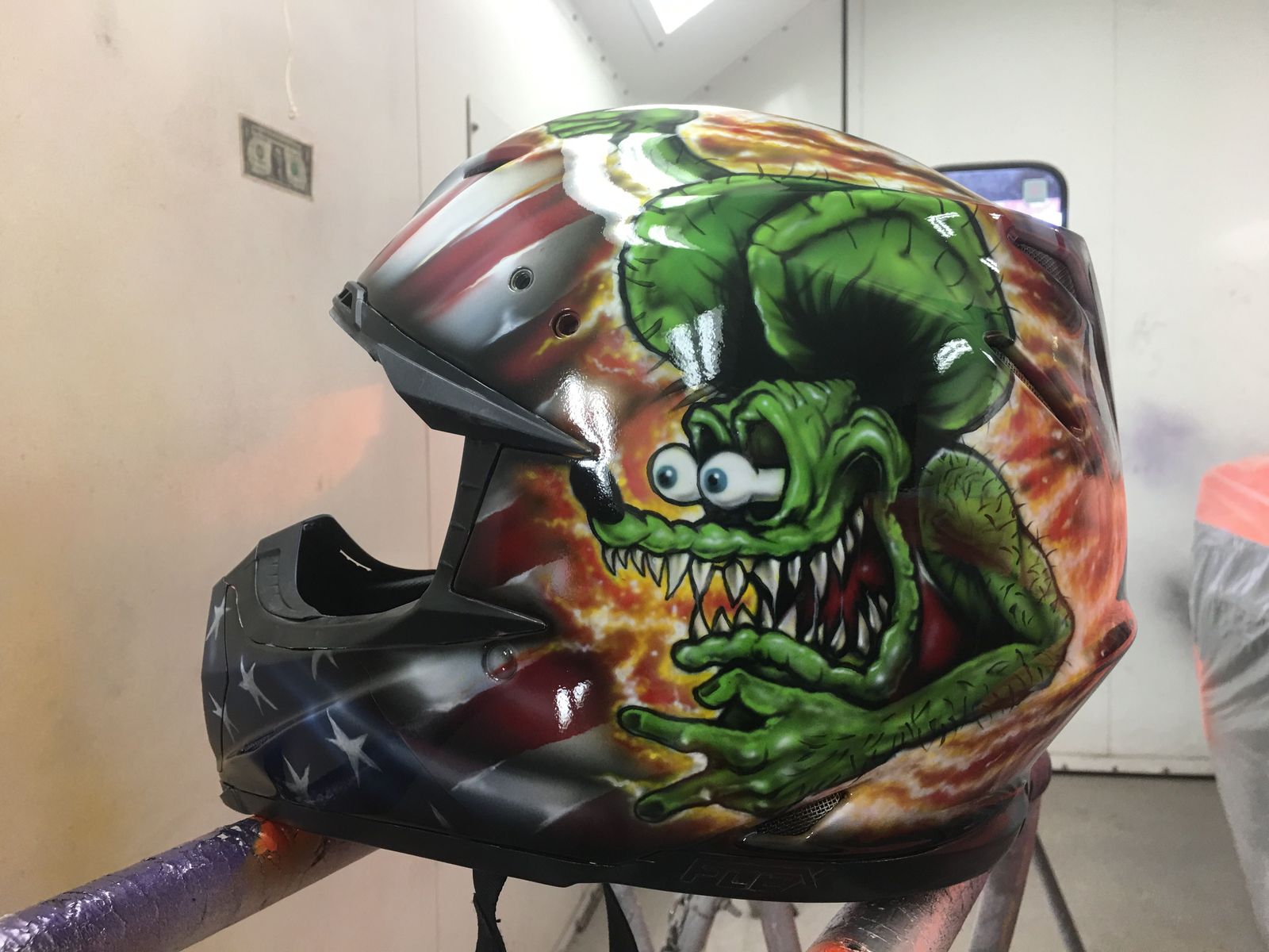 A motocross helmet in a paint booth airbrushed with a bug-eyed green monster over an American flag and orange flames.