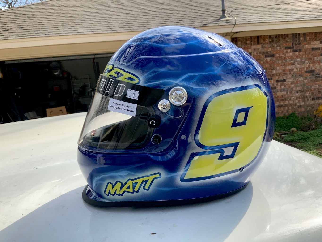 A full-face racing helmet airbrushed in blue with white lightning, a large yellow number 9 and a first name in yellow script, sitting on a white car roof.