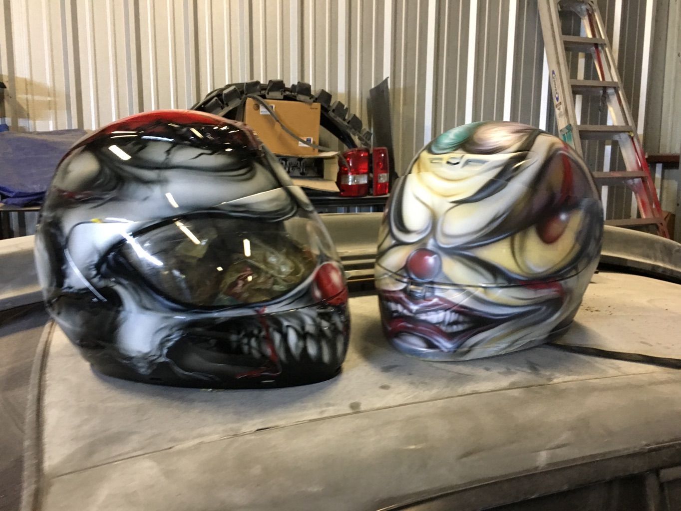 Two full-face motorcycle helmets on a workbench, one airbrushed as a grinning grey skull, the other as a pale clown face with a red nose.