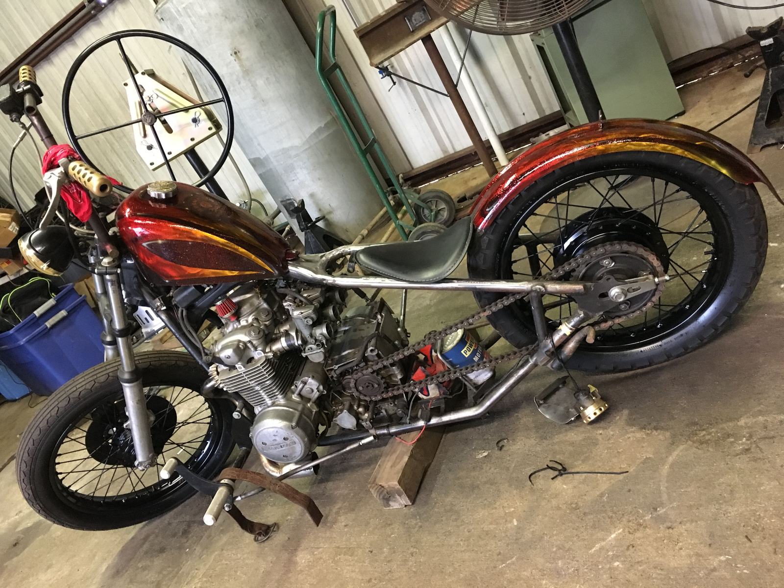 A stripped-down chopper motorcycle in a workshop, its teardrop tank and rear fender painted deep red fading to gold.