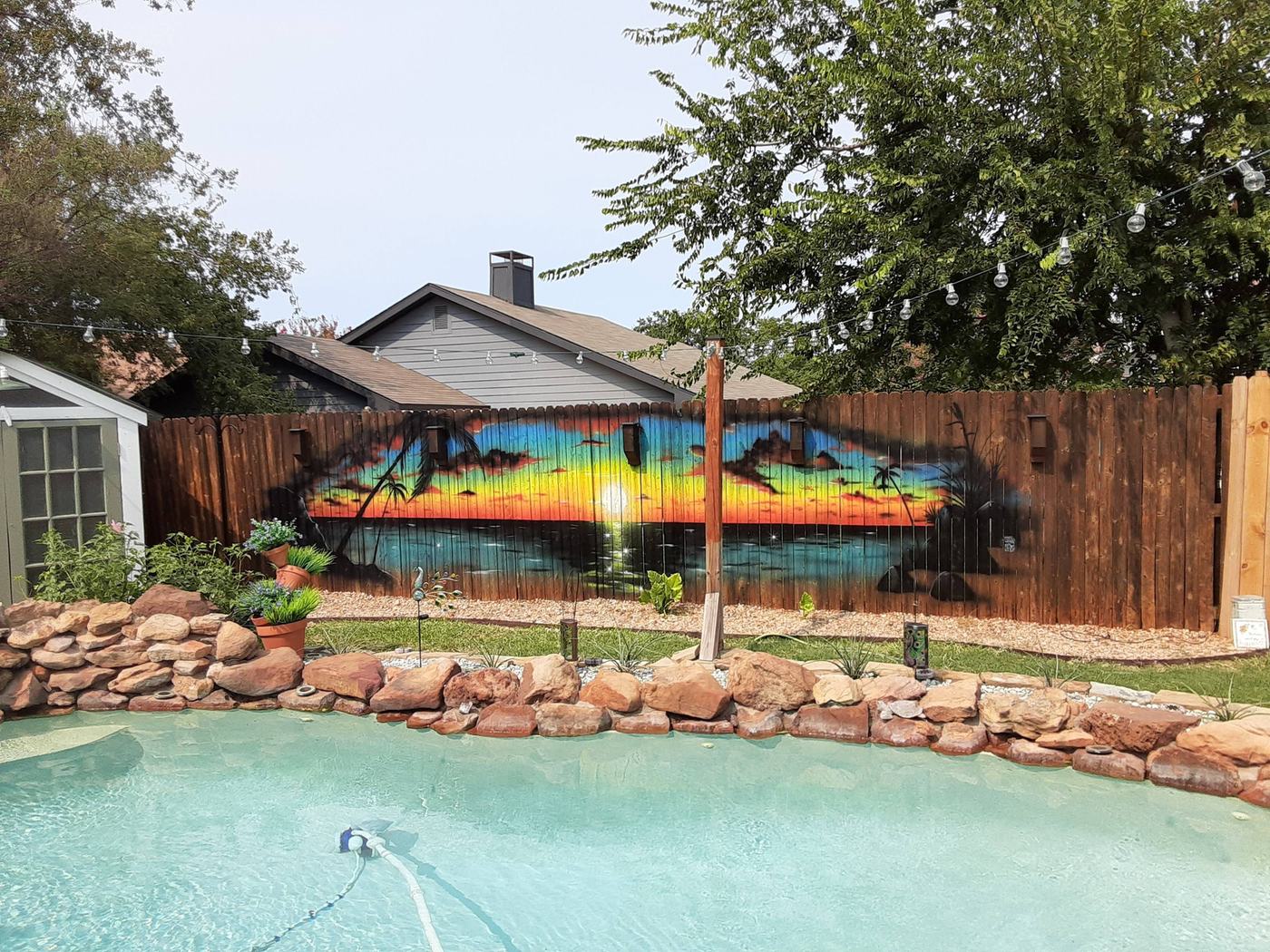 A wooden backyard fence behind a swimming pool and rock border, painted with a bright tropical sunset, palm trees and silhouetted islands.