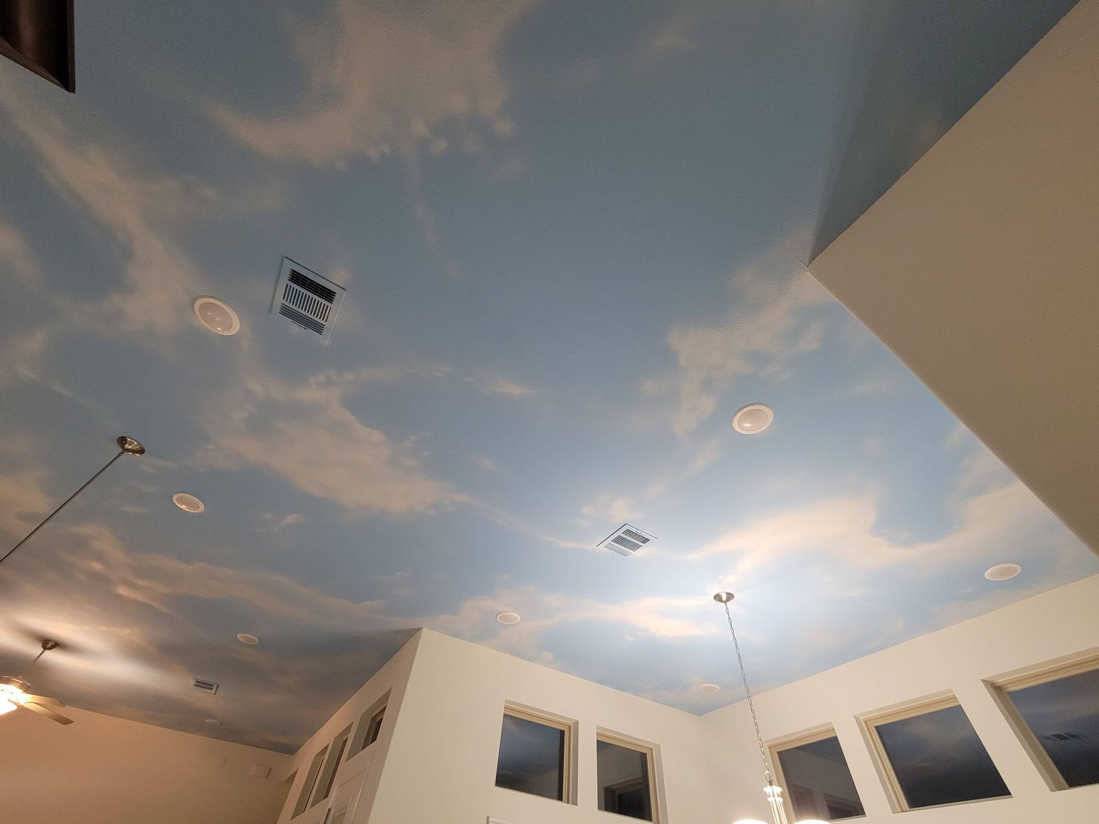 A high ceiling painted as a pale blue sky with drifting clouds, seen from below, with recessed lights, air vents and a row of upper windows.