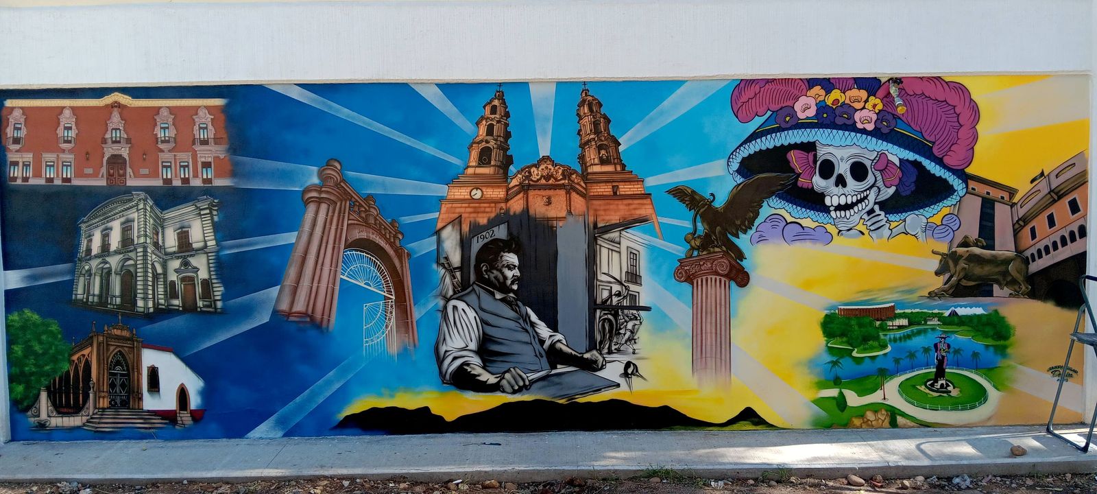 A long exterior wall mural painted with church towers, a stone archway, a skull-faced Catrina in a flowered hat, an eagle and a seated man at a desk, against blue sunburst rays.