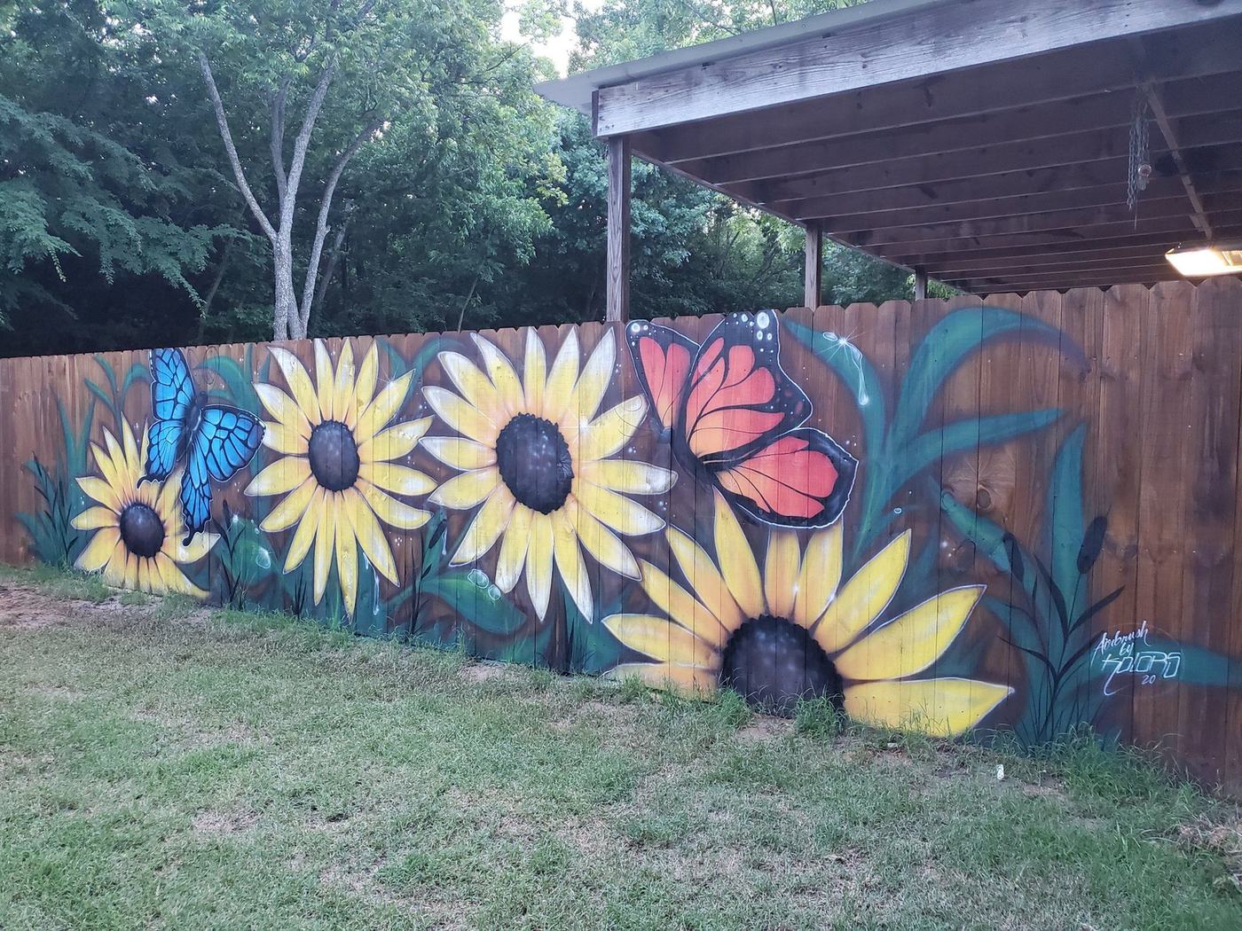 A long wooden fence in a grassy yard painted with large yellow sunflowers, a blue butterfly and an orange butterfly.