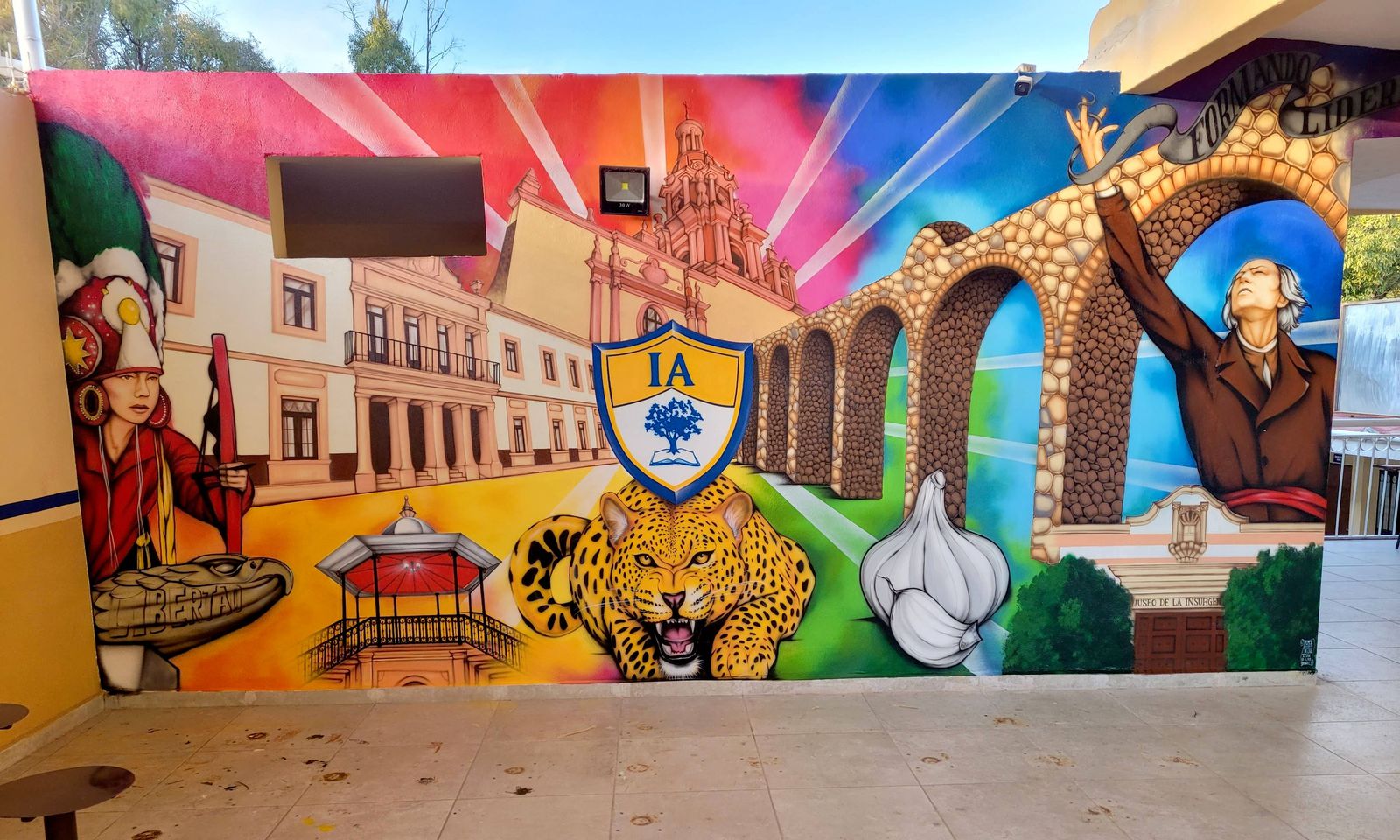 A long, brightly coloured mural on a covered patio wall showing colonial buildings, a stone aqueduct, a jaguar, a garlic bulb, a crest with the letters IA and a historical figure with a raised arm, under rainbow sun rays.