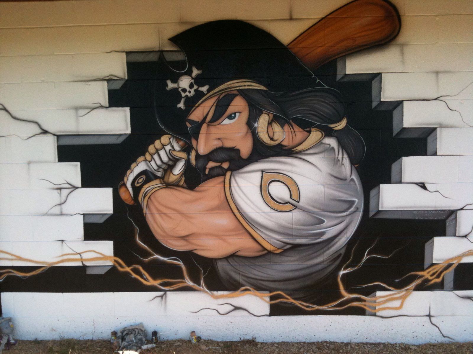 An exterior block wall painted with a muscular pirate mascot in an eye patch and hat, gripping a baseball bat and breaking out through painted cinder blocks.