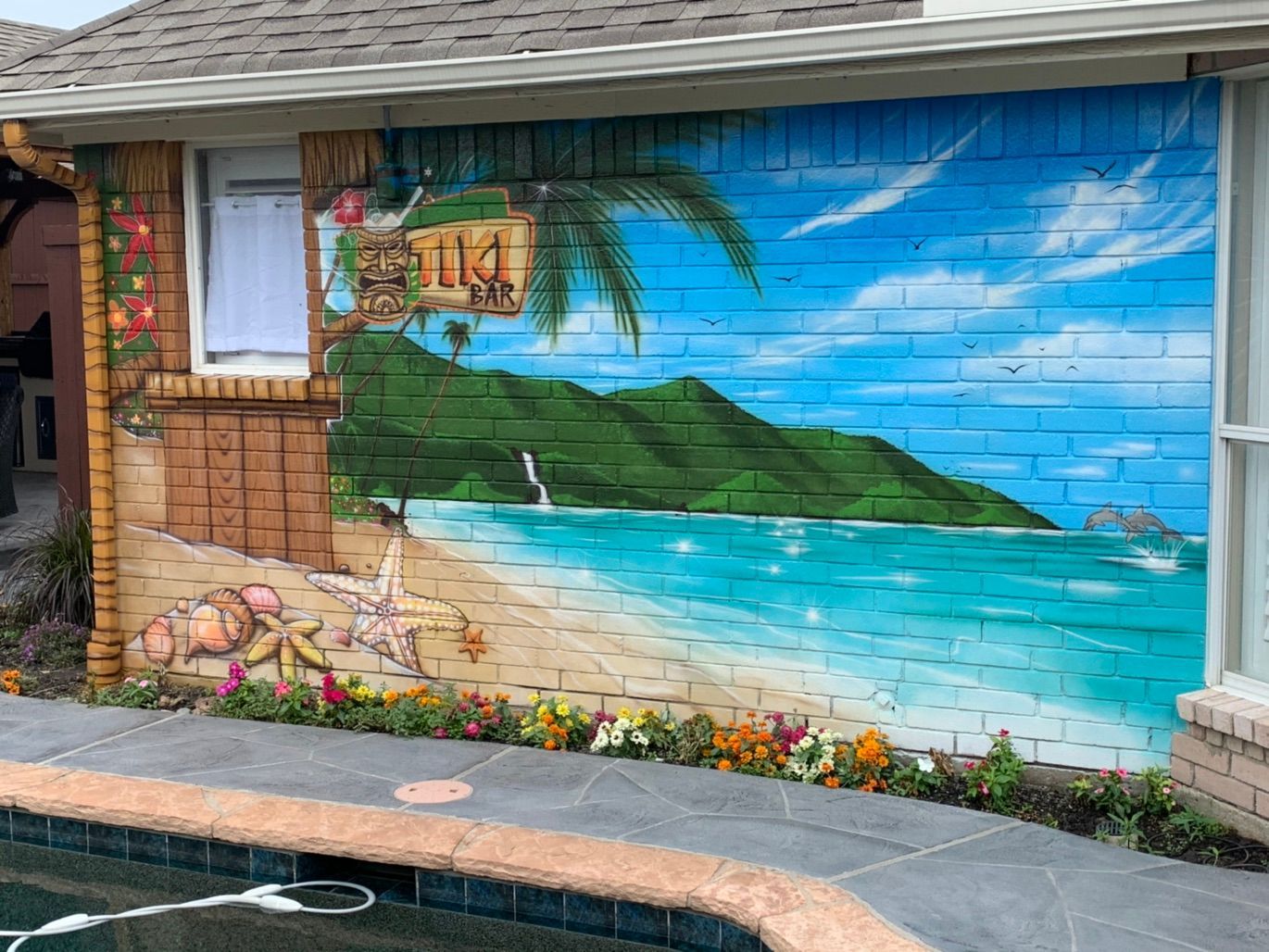 An exterior brick wall beside a pool painted with a tiki bar sign, a sandy beach with shells and a starfish, turquoise water and green mountains.