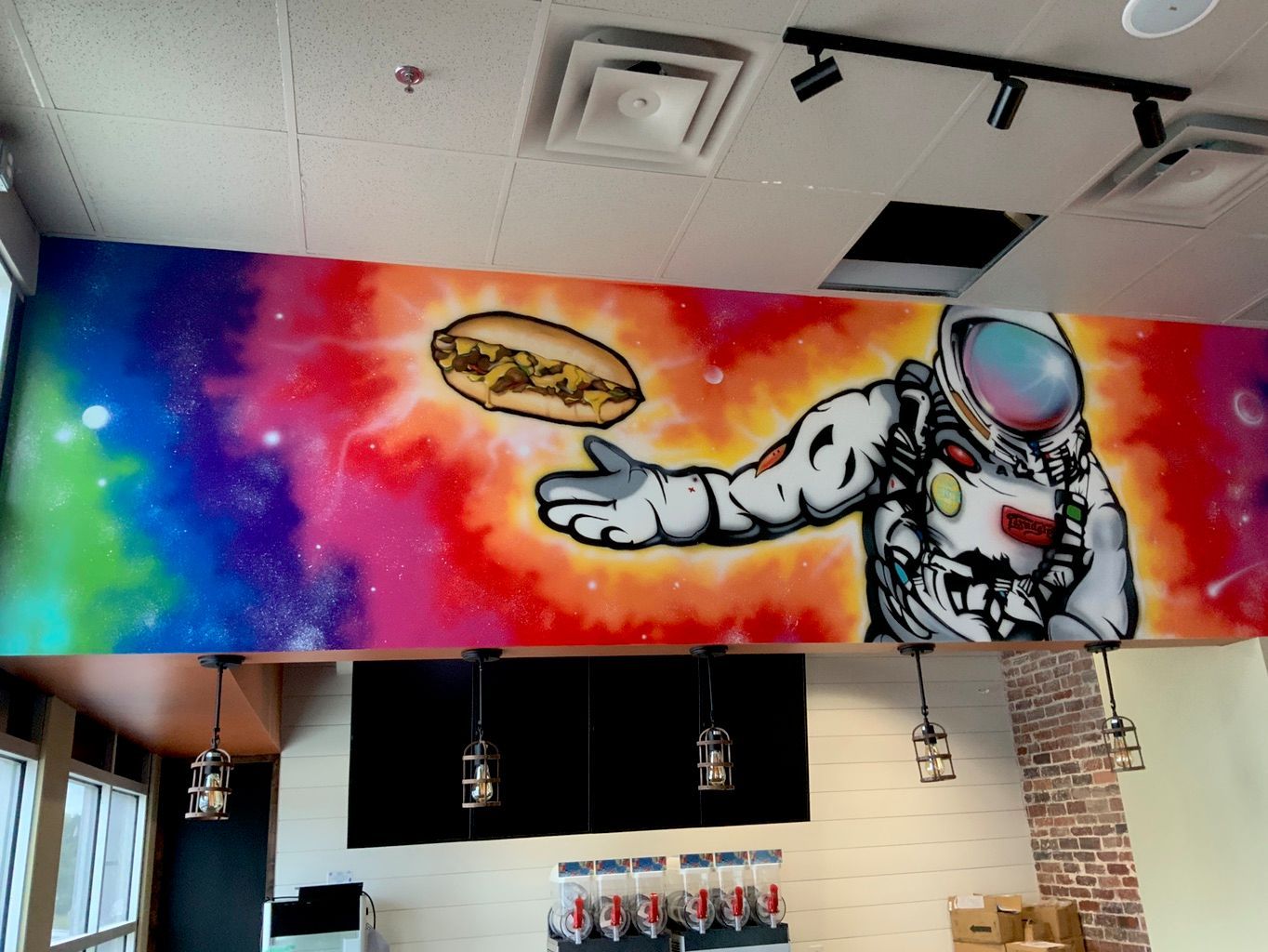 A long restaurant wall above the counter painted with a rainbow galaxy and an astronaut in a white suit reaching toward a floating sandwich.