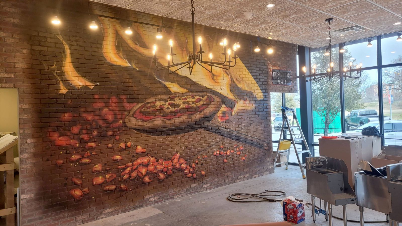 Inside a restaurant being fitted out, a brick wall painted to look like a glowing wood-fired oven, with flames, embers and a pizza on a peel, under hanging chandeliers.