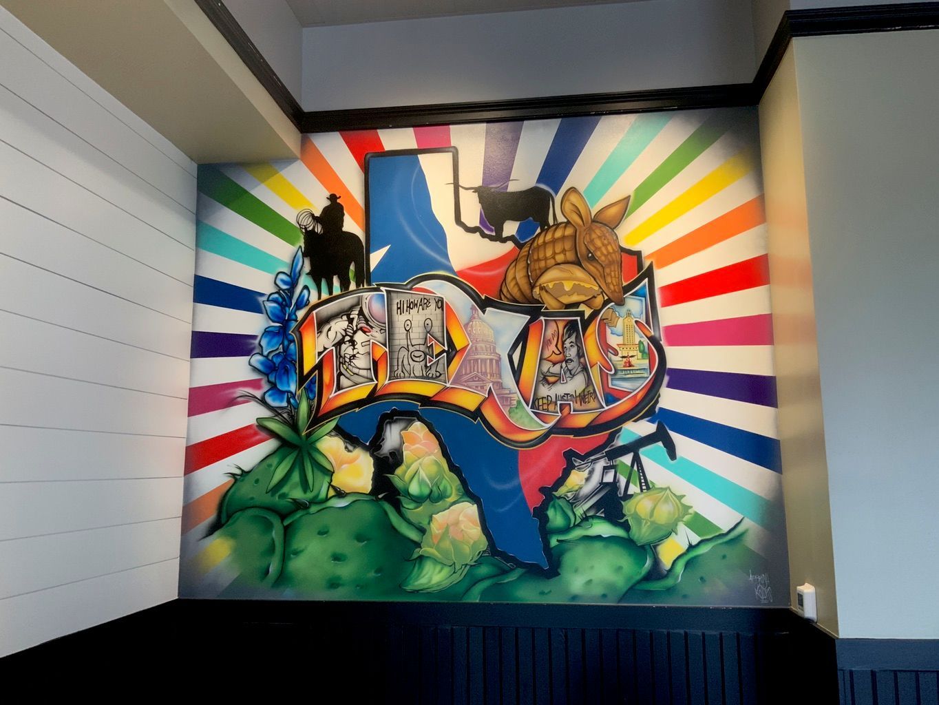 A restaurant wall painted with rainbow sunburst rays behind a map of Texas, the word TEXAS in graffiti letters, an armadillo, bluebonnets, a cowboy on horseback and prickly pear.