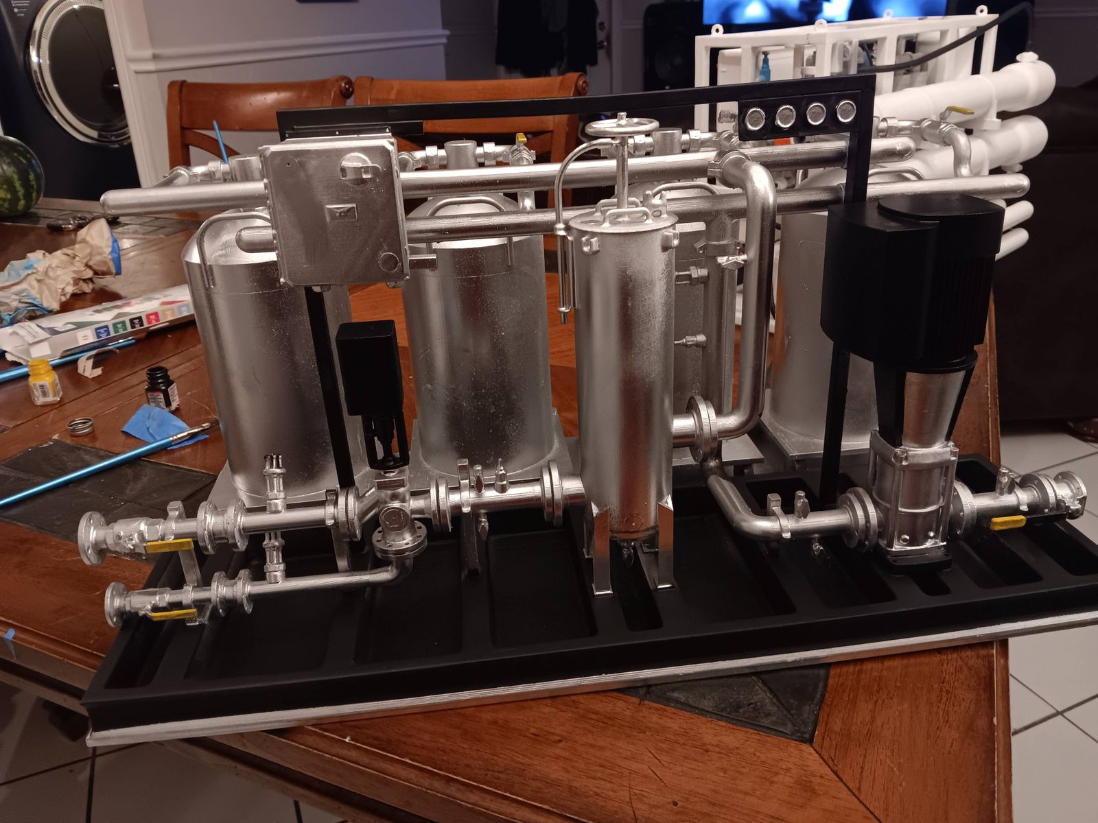 A detailed scale model of industrial process equipment, with tanks, pipes and valves painted metallic silver and black, on a table.