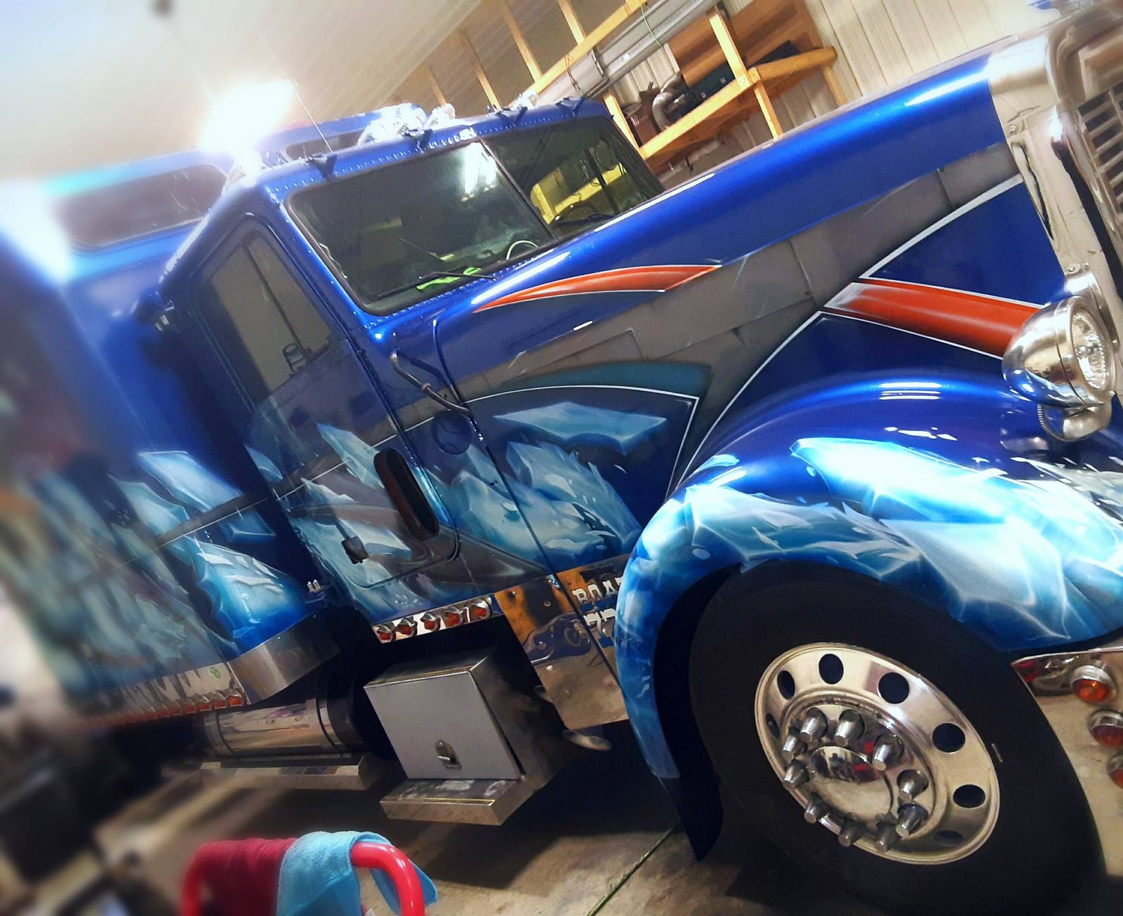 A long-nose semi truck in a workshop, painted deep metallic blue with airbrushed water and ice effects across the hood, fenders and cab, and an orange stripe along the hood.