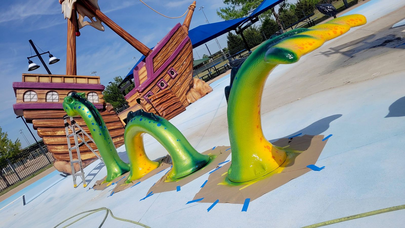 Three arching sea-serpent segments at a splash pad painted in green and yellow, with a wooden pirate ship play structure behind them.