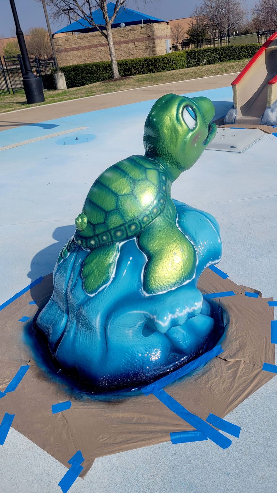 A sea turtle sculpture at a splash pad, freshly painted in glossy greens on a blue wave-shaped base, with masking paper still taped to the ground around it.