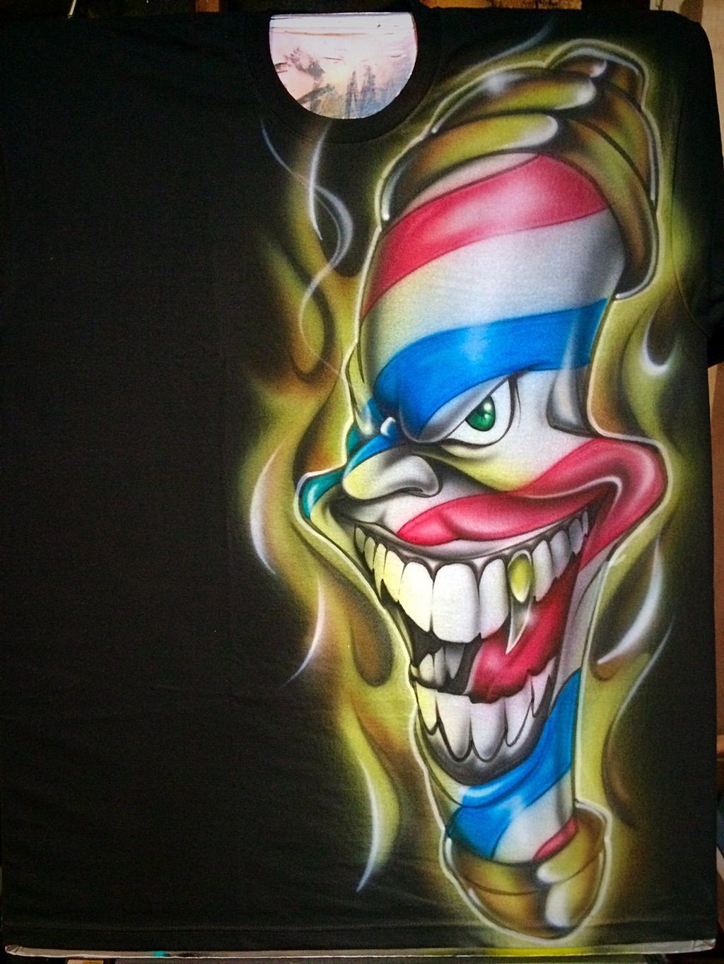 A black T-shirt airbrushed with a grinning, clown-faced barber pole in red, white and blue, wreathed in yellow smoke.