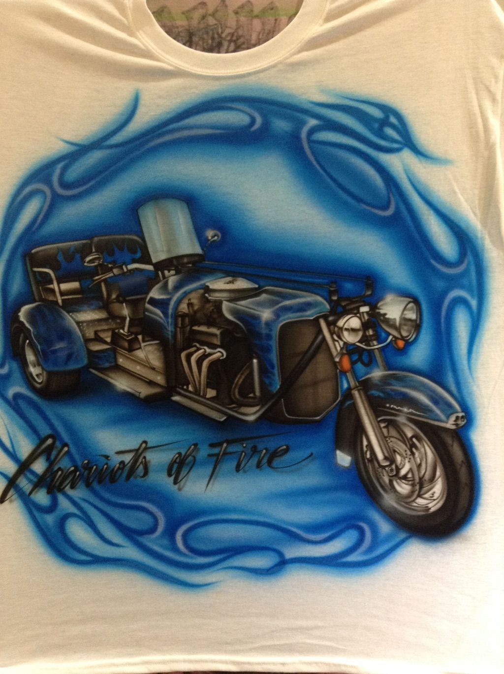 A cream T-shirt airbrushed with a motor trike framed by blue tribal flames, with script lettering beneath.