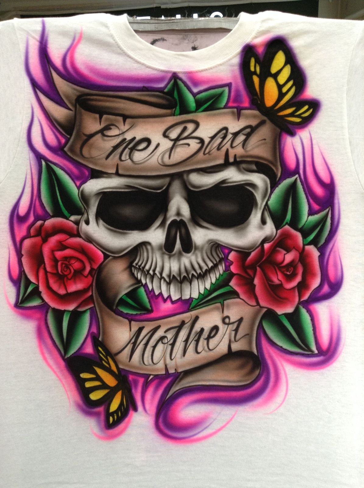 A white T-shirt airbrushed with a skull between red roses and yellow butterflies, wrapped in a banner with script lettering, on pink and purple flames.